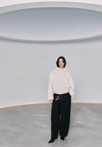 Persoană purtând un hanorac alb pufos și pantaloni largi negri, stând pe podeaua gri sub o lumină circulară mare de plafon.