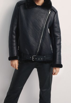 Person mit einer schwarzen Fliegerjacke aus Kunstlammfell mit asymmetrischem Reißverschluss, breitem Kragen, Gürtel am Saum und passenden schwarzen Hosen.