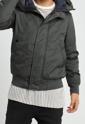 Mand iført en mørkegrå hættetrøje-bomberjakke over en hvid og sort stribet skjorte, med sorte bukser, poserer med den ene hånd nær sit hoved.