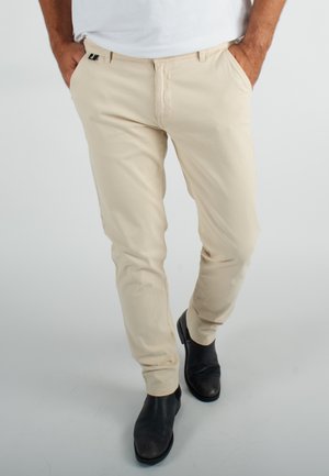 Pantalons chino beiges en tissu léger avec une coupe slim, dotés de poches latérales et d'un petit logo accentué sur la ceinture. Bottes noires incluses.