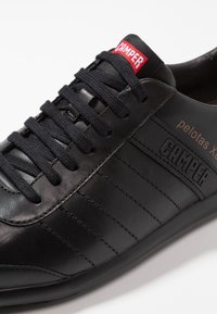 Black leather sneakers with a smooth texture, featuring horizontal stitching, black laces, and a red brand label on the tongue.