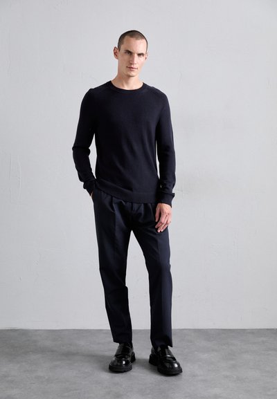 Homme debout contre un mur pâle, portant un pull noir, un pantalon sombre et des chaussures en cuir noir, avec une main dans sa poche.