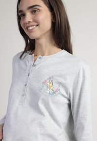 Camisa de manga larga gris claro con un cuello abotonado y un diseño bordado que presenta un personaje rodeado de flores y texto.