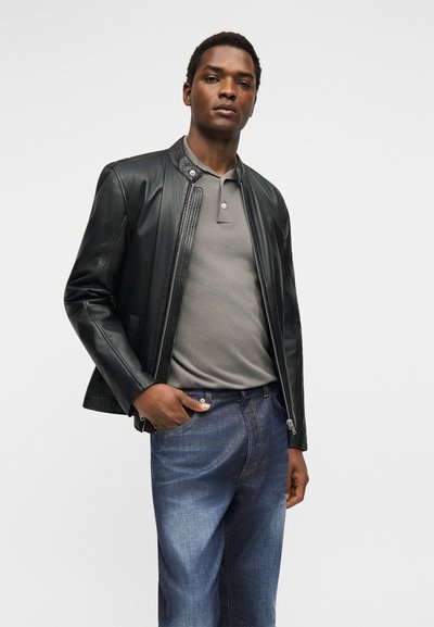 Άνδρας με μαύρο δερμάτινο μπουφάν πάνω από γκρι πόλο μπλουζάκι και μπλε τζιν, στέκεται με το ένα χέρι στην τσέπη μπροστά σε λευκό φόντο.