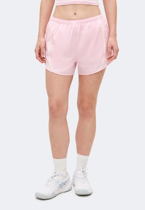 Γυναίκα που φοράει ανοιχτό ροζ αθλητικό σορτς, λευκές κάλτσες και λευκά παπούτσια για τρέξιμο, όρθια με τα πόδια ελαφρώς ανοιχτά σε απλό φόντο.