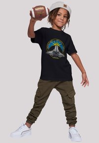 Black t-shirt features a graphic of a space shuttle and sunburst. Paired with olive cargo pants and white sneakers, holding a football.