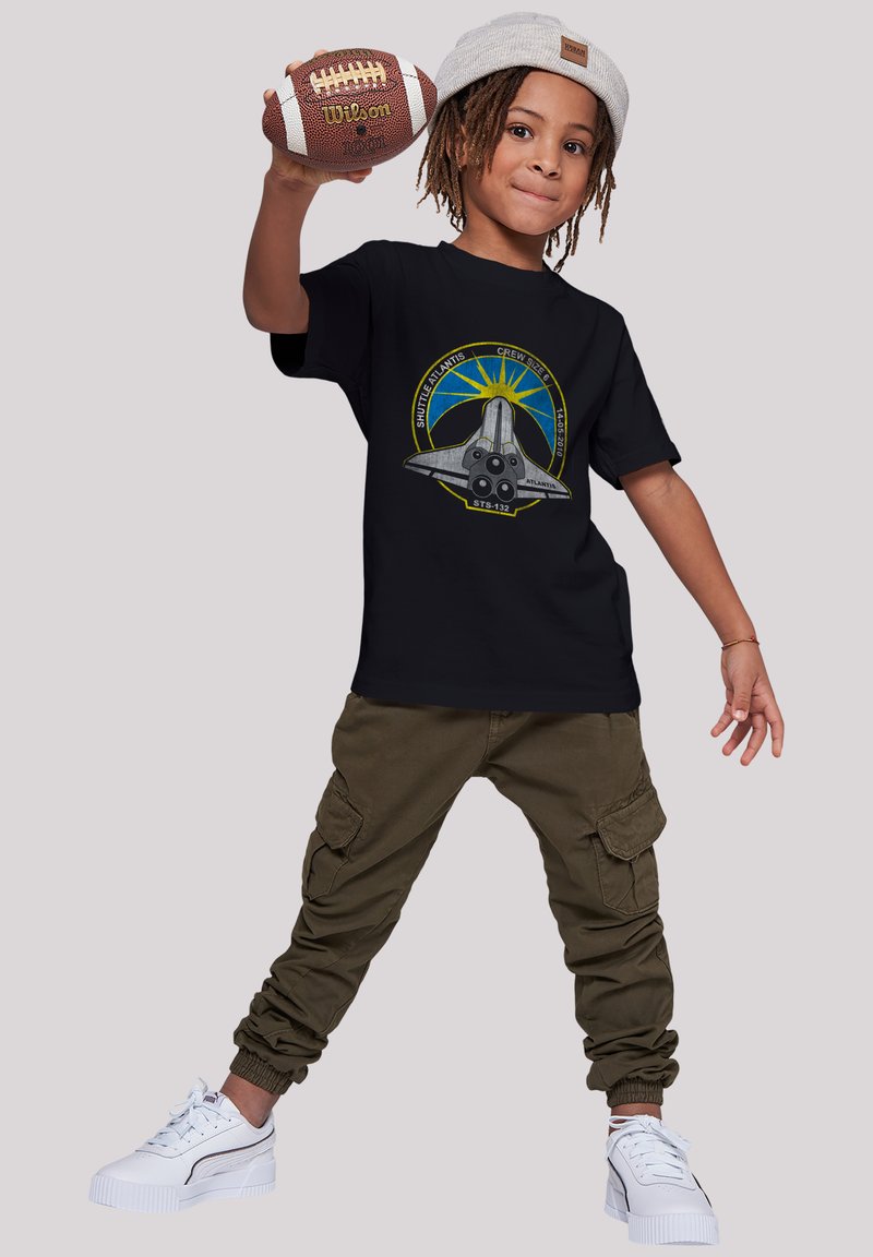 Black t-shirt features a graphic of a space shuttle and sunburst. Paired with olive cargo pants and white sneakers, holding a football.