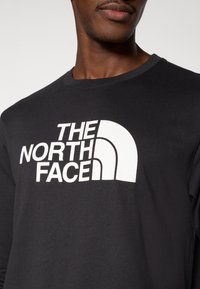 Μαύρο μακρυμάνικο μπλουζάκι με εξέχον λευκό λογότυπο, "THE NORTH FACE", σε έντονη, καθαρή τυπογραφία στο μπροστινό μ έρος.