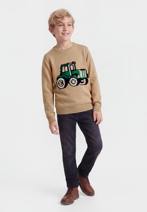 Garçon blond portant un pull beige avec un motif de tracteur vert, jean noir et bottes marron, debout et souriant sur fond blanc.