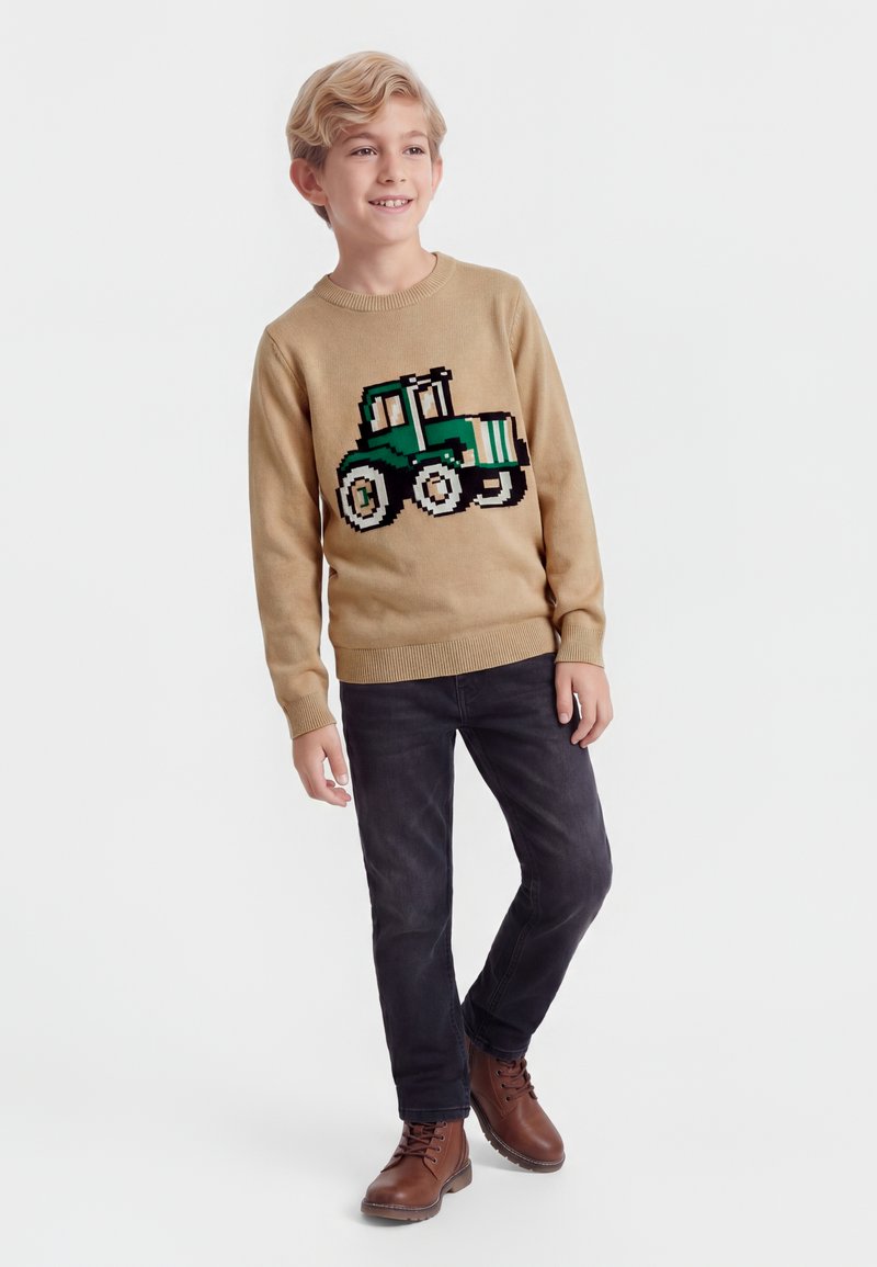 Garçon blond portant un pull beige avec un motif de tracteur vert, jean noir et bottes marron, debout et souriant sur fond blanc.