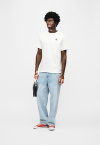 Λευκό βαμβακερό t-shirt με μικρό μαύρο λογότυπο, ανοιχτό μπλε τζιν παντελόνι με φαρδιά ποδιά και κόκκινα και λευκά αθλητικά παπούτσια. Κρατάει μαύρη τσάντα με ασημένιες λεπτομέρειες.