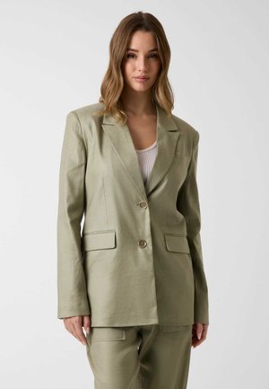 Joven mujer con cabello ondulado hasta los hombros, que lleva un blazer verde oliva claro y pantalones a juego sobre una camiseta blanca acanalada, de pie frente a un fondo gris.