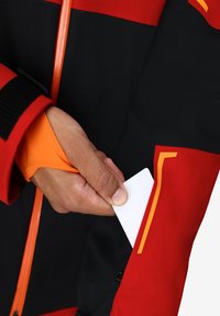 Giacca rossa e nera con accenti arancioni; presenta una tasca con zip che contiene una scheda bianca. Il materiale sembra resistente con una finitura liscia.