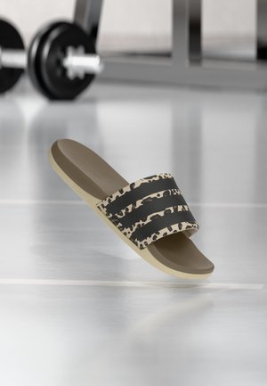 Beige slidssandal med sorte striber og leopardprintrem på skinnende gulv, træningsbænke og håndvægt i baggrunden.
