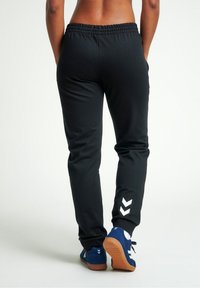 Schwarze Sporthosen mit einem elastischen Bund und einem weißen Chevron-Logo am unteren linken Bein, kombiniert mit blauen Laufschuhen.