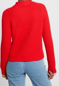 Červený pletený svetr s žebrovanou strukturou, raglánovými rukávy a kulatým výstřihem; kombinovaný se světle modrými džínami.