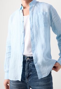 Camicia button-up in lino blu chiaro con maniche lunghe, taschino sul petto e vestibilità casual, abbinata a un paio di pantaloni in denim blu.