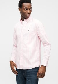 Άνδρας που φοράει πουκάμισο με ριγέ σε απαλό ροζ και άσπρο, με μικρό λογότυπο στο στήθος, συνδυασμένο με σκούρο μπλε τζιν, κοιτάζοντας προς το πλάι.