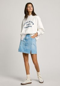Sudadera blanca con texto azul marino, combinada con una falda de mezclilla azul clara con una abertura frontal. La modelo lleva zapatillas blancas y calcetines claros.