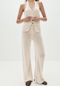 Beige stickad halterneck-topp med framknappar, parat med matchande vida byxor. Tyget är lätt och genomskinligt, med snörningsdetalj.