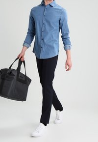 Ljusblå denimskjorta med knappar, mörka byxor, vita sneakers och en svart strukturerad handväska. Man som går avslappnat.