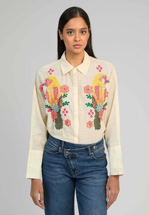 Femme portant une chemise crème brodée de perroquets colorés et de fleurs, associée à un jean bleu, sur un fond uni gris clair.