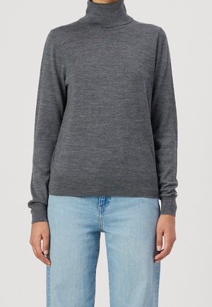 Persona che indossa un maglione aderente a collo alto grigio scuro con maniche lunghe e jeans di denim azzurro chiaro su uno sfondo semplice.