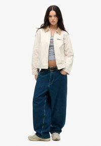 Hellbeige Canvas-Jacke mit braunem Kragen, Reißverschlusstasche und lockeren blauen Denim-Hosen. Lässige Schuhe runden das Outfit ab.