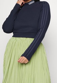 Marinblå ribbad långärmad crop top med hög hals och tre mörka ränder på ärmarna, som matchas med en plisserad ljusgrön kjol.