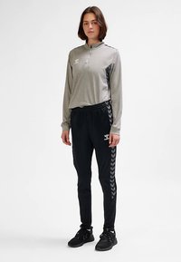 Hellgraues Langarmoberteil mit Reißverschlusskragen und schwarzen Akzenten, kombiniert mit schwarzen, zulaufenden Hosen mit Chevron-Muster entlang der Seiten.