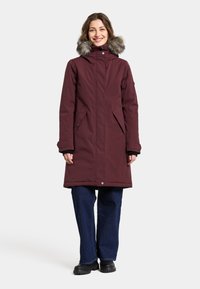 Bordeauxfarbener wasserdichter Parka mit Kapuze aus Kunstpelz, Seitentaschen und einem mid-length Design. Kombiniert mit weiten dunkelblauen Jeans und schwarzen Stiefeln.