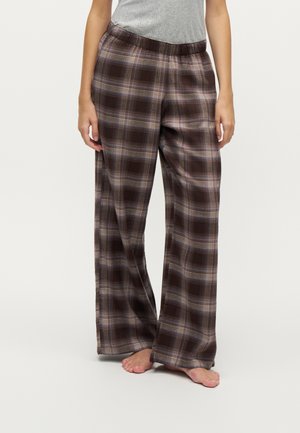 Persona che indossa pantaloni larghi a quadri marroni e un top grigio senza maniche, in piedi a piedi nudi contro uno sfondo semplice.