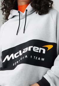 Szara bluza z kapturem z czarnym panelem z napisem "McLaren" oraz "Formula 1 Team" na białym tle. Czarny sznurek do ściągania, ribbed mankiety oraz pomarańczowy wewnętrzny kołnierz.