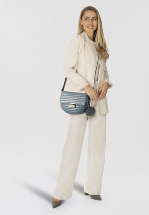 Borsa a tracolla in materiale blu testurizzato con design trapuntato, dotata di patta anteriore e hardware metallico. Indossata con un completo crema e tacchi.