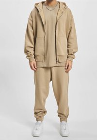 Beiger Baumwoll-Trainingsanzug mit einem Kapuzenpullover mit Reißverschluss, großen Taschen, langen Ärmeln und passenden, schmal zulaufenden Hosen. Kombiniert mit weißen Turnschuhen.