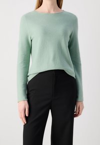 Frau trägt einen hellgrünen langärmligen Stricktop, in eine hoch taillierte schwarze Hose gesteckt, und steht vor einem schlichten weißen Hintergrund.