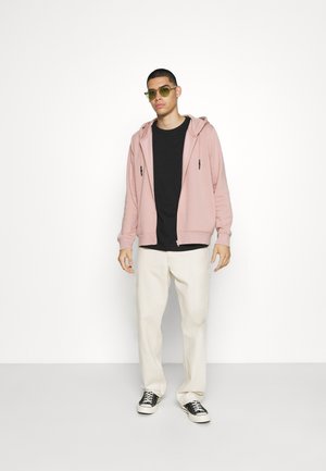Svetlo roza zip-hoodie preko črne majice, v kombinaciji s smetanovimi hlačami in črnimi teniskami. Dodatki vključujejo zelene sončne očala.