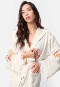 Biely textúrovaný župan s koralovým vzorom vyšitým v oranžovej a červenej farbe na rukávoch, s kapucňou, strapcovými manžetami a viazacími pásmi.