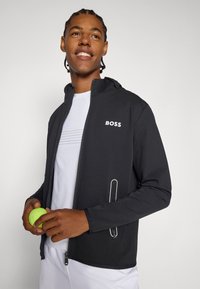 Svart zip-up jacka i stretchmaterial, med subtil logotyp, sidofickor med dragkedjor och en huva, bärs över en vit skjorta.