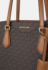 Bruine en donkergepatronde Michael Kors handtas met dubbele leren handvatten en een gouden logo plaat centraal nabij de bovenrand.