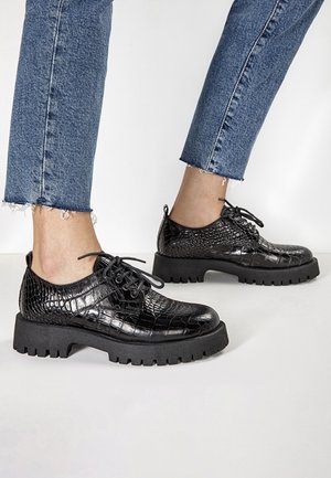 Czarne sznurowane buty z croco-teksturowanej skóry, masywna czarna gumowa podeszwa i zaokrąglony czubek. Połączone z jeansami w niebieskim kolorze z postrzępionymi nogawkami.