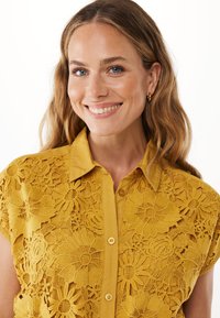 Camicia gialla senape con dettaglio in pizzo floreale, Maniche corte, chiusura con bottoni davanti e tessuto liscio con motivi intricati.