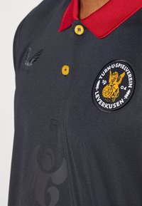 Donkergrijze poloshirt met een rode kraag, voorzien van gele knopen, een embleem met een gevleugeld figuur en subtiele textuurdetails.