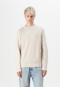 Light beige sweater with a textured knit pattern, round neck, and ribbed cuffs. Paired with light blue denim jeans.