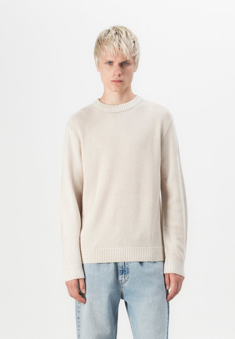 Light beige sweater with a textured knit pattern, round neck, and ribbed cuffs. Paired with light blue denim jeans.