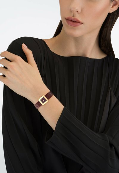 Burgundy bőr óraszíj aranyszínű négyzet alakú tok és sötét számlap. Fekete, redőzött felső széles ujjakkal a modell karján.