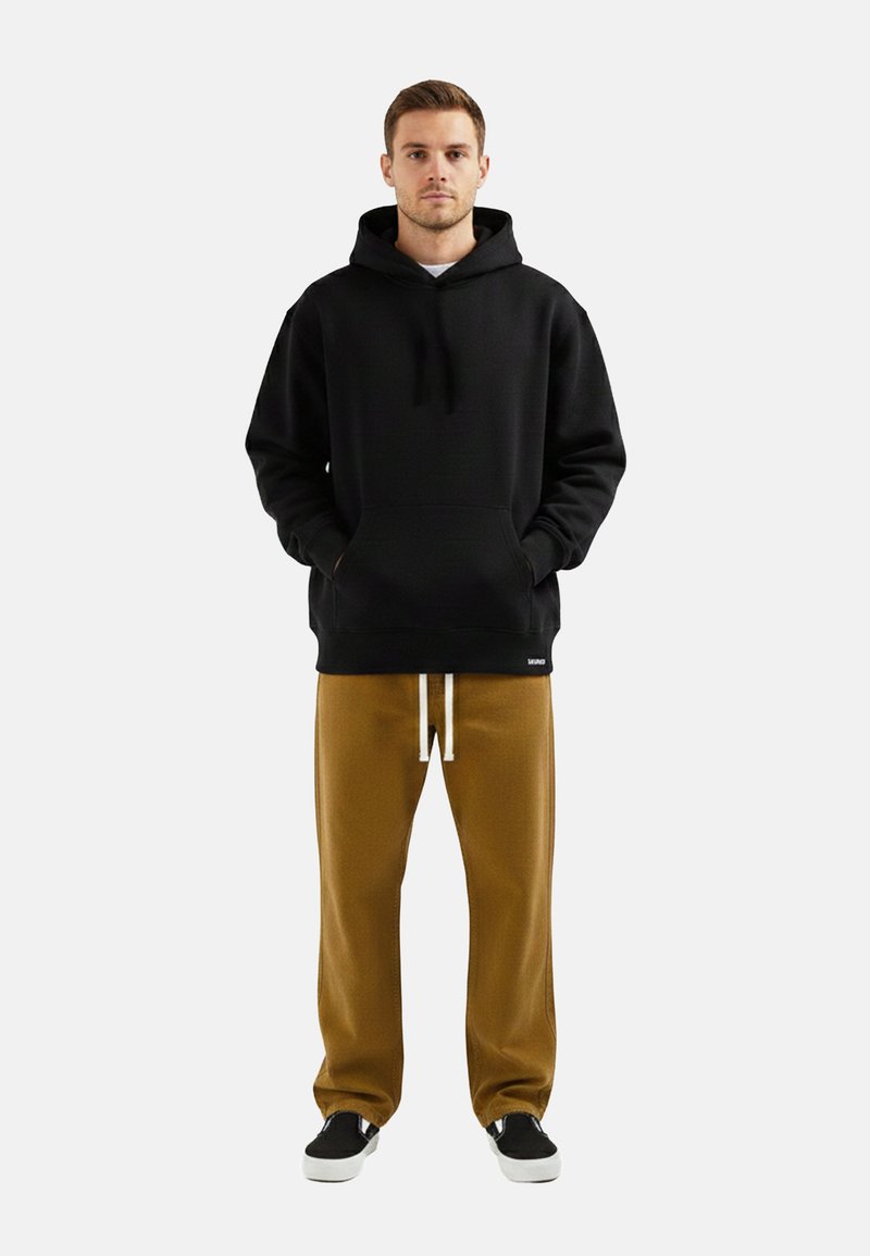 Un homme se tient face à l'objectif, vêtu d'un sweat à capuche noir, d'un pantalon de survêtement marron avec des cordons blancs, et de chaussures noires à enfiler avec des semelles blanches.