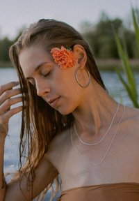 Silver hoop earrings, layered silver necklaces, and a coral flower accentuate wet hair against a lake background. Brown strapless top visible.