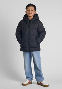 Giacca a vento nera con zip frontale e cappuccio, abbinata a jeans azzurri chiari e scarpe bianche con accenti rossi.