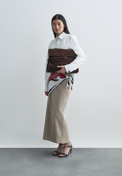 Frau in weißem Hemd, beiger Rock und schwarzen Absatzsandalen hält eine große braune Clutch-Tasche mit Gürtel-Detail.